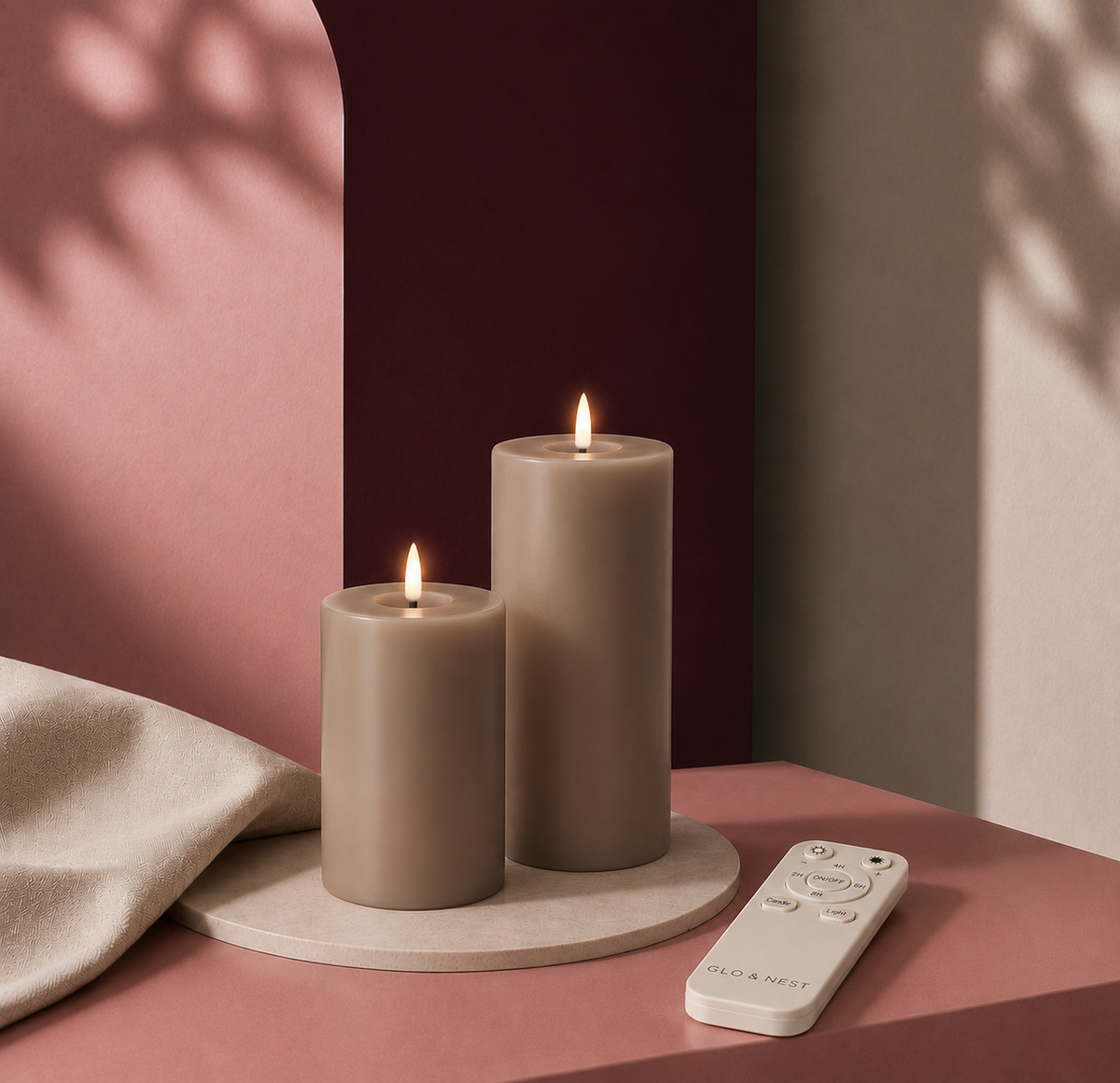 Two lit matte pillar candles in warm taupe tones sit on a round stone coaster on a pink table, with a white remote control nearby and a draped beige cloth against a pink and dark burgundy backdrop.